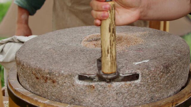 Close up of baker rotating wooden handle on traditional medieval stone hand mill, grinding grain into freshly ground coarse whole grain flour.