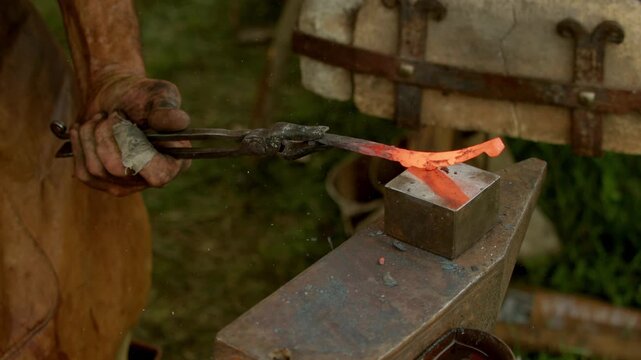 Macro closeup shot captures strong hands of medieval blacksmith hammering a glowing red-hot steel workpiece held with tongs on a heavy iron anvil in traditional outdoor forge...