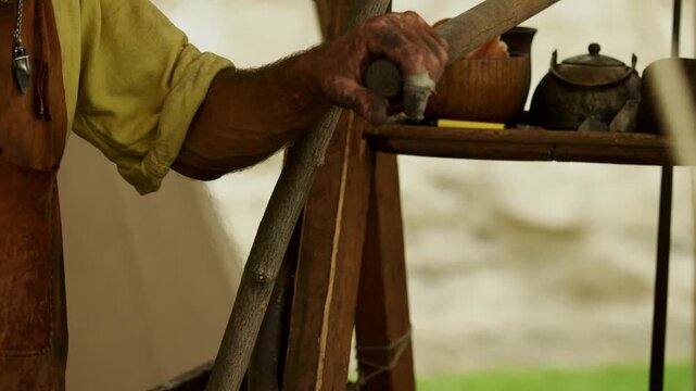 Closeup shot captures soot-covered hand of medieval blacksmith operating a wooden lever to pump air bellows, blowing airflow and sparks into hearth fire.