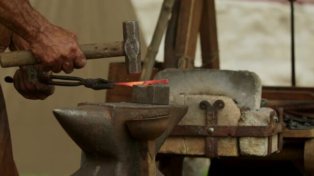 Medium closeup shot captures medieval blacksmith striking a glowing red-hot steel bar with a heavy hammer on an iron anvil beside a portable stone forge hearth in an open-air workshop...