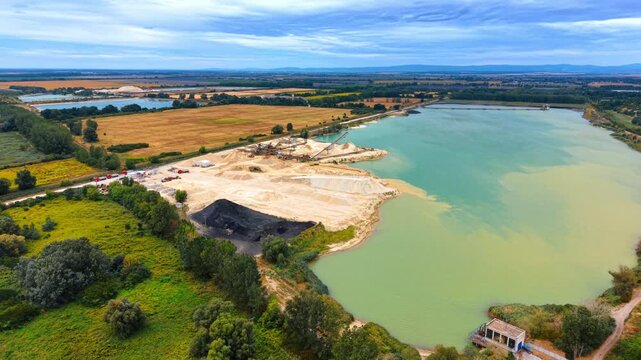 Wide aerial view of open pit sand quarry and lake. Green trees and cultivated farmland envelope the industrial mining site and clear water pond.