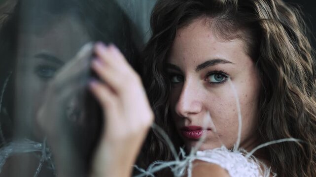 Young woman with seductive eye look at camera while leaning against a reflective glass surface with reflection, slow motion
