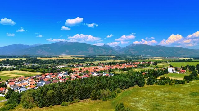 Village in valley below mountain range. Red roofs line green fields near a forest with mountains on the horizon.
