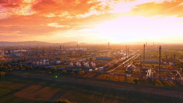 Panoramic view of oil refinery complex under orange sky. Industrial zone with distillation units, agricultural fields, bright horizon, evening glow.
