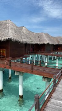 Vertical video of wooden luxury overwater bungalows with thatched roofs built on stilts over clear turquoise ocean water connected by a wooden boardwalk in the Maldives.