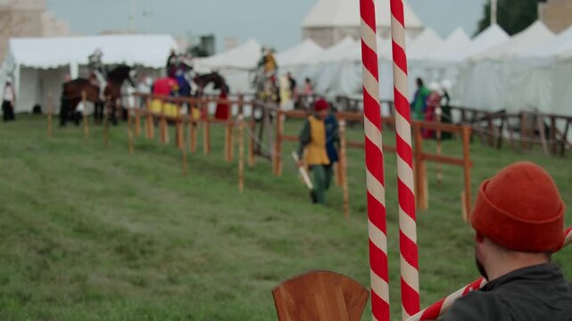 Medium shot captures red and white spiral-striped jousting lances held in foreground as medieval tournament arena prepares with mounted knights, squire, and white camp tents in background.