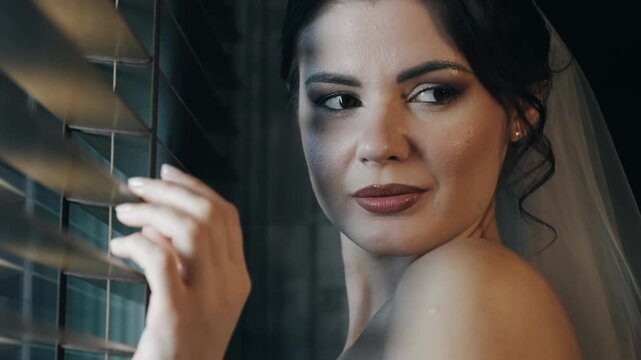Portrait of excited seductive bride woman standing in a thoughtful pose near window blinds looking at camera with teasing eyes