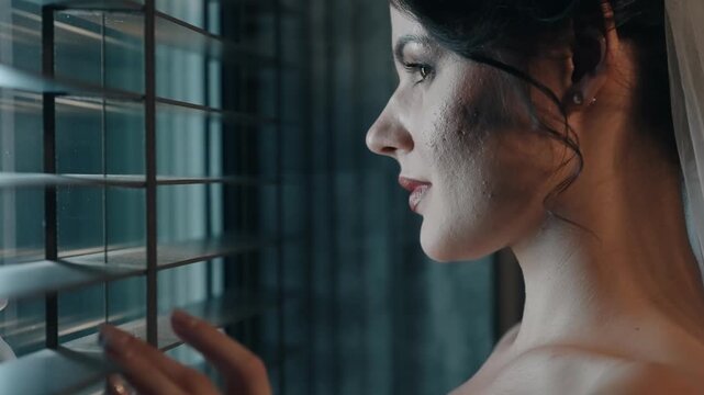 Young bride woman looking thoughtfully through window blinds at night with her face illuminated gently