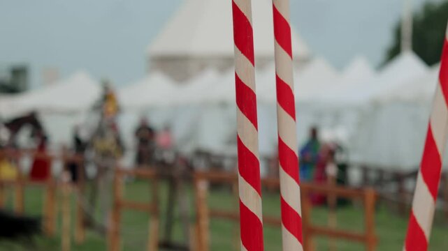 Medium closeup shot focuses on red and white striped tournament lances held by squire while mounted knight in armor on horseback appears blurred in background.