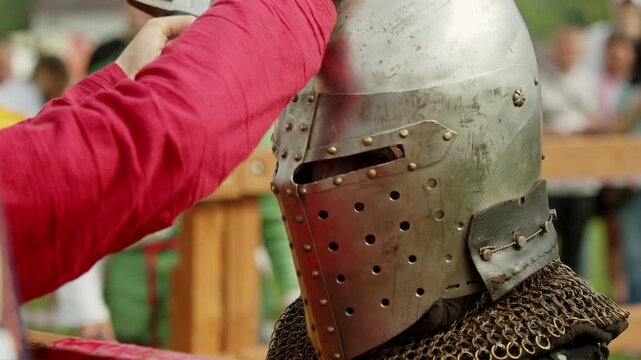 Closeup shot captures squire in red tunic striking and fixing medieval knight's steel bascinet helmet visor using a heavy sword pommel during tournament pause.