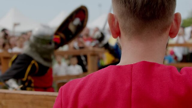 Man in red medieval tunic watching two heavily armored knights aggressively clashing with swords and shields in tournament arena.