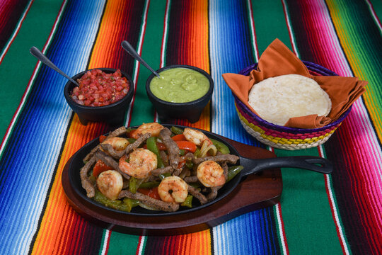Sizzling beef and shrimp fajitas with saut&eacute;ed bell peppers and onions, served in a cast-iron skillet alongside flour tortillas, fresh tomato salsa, and creamy guacamole on a colorful Mexican-style str