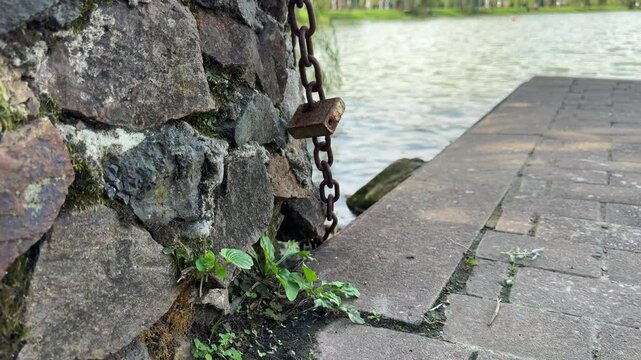 Old rusty metal chain with padlock hangs on stone wall near paved pier on lake shore in public park on sunny day
