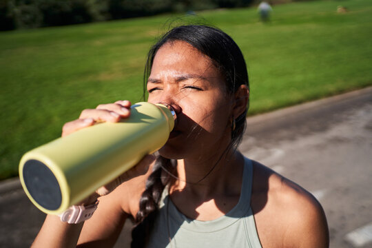 Black Latina woman drinking from a reusable water bottle after exercising in a sunny park. Close-up fitness scene conveying hydration, recovery and an active healthy lifestyle.