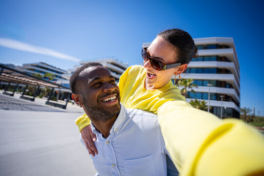 Cheerful interracial couple laugh closely together during a piggyback pose in a modern residential area under clear sunny blue sky.