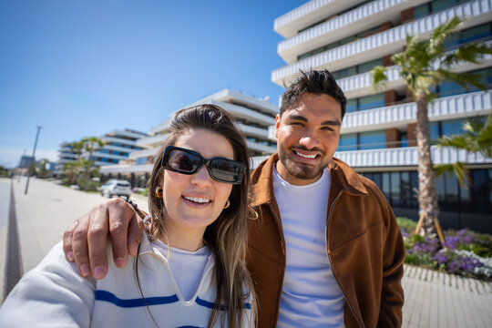 Smiling young couple stand close together for a selfie in a contemporary residential district with blue sky, palm trees and bright sunshine.