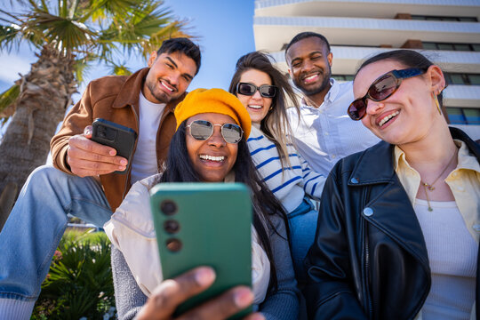 Multicultural friends sit closely together and smile while sharing content on a smartphone in a sunny modern residential district with palm tree and blue sky.