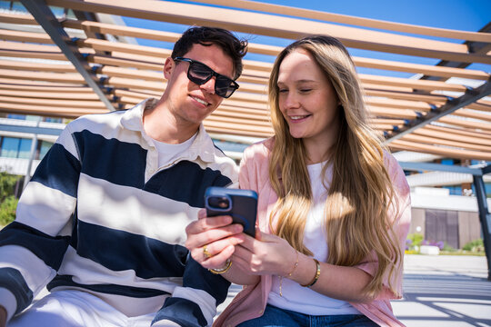 Young couple sit closely together under a shaded pergola, smiling while looking at a smartphone and sharing digital content during a relaxed sunny day.