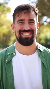 Vertical. Cheerful Caucasian millennial man standing in an autumn park and smiling directly at the camera. Outdoor portrait with natural light and blurred background conveying confidence and friendly
