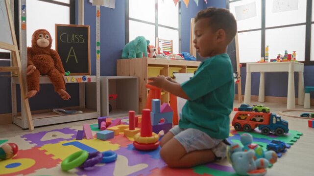 Boy stacking colorful toy rings and kneeling on foam playmat in classroom building, reaching with hand toward blocks and toy truck; curiosity learning play.