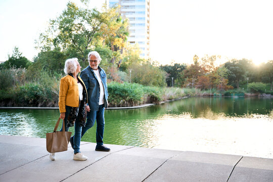 Senior couple walking and talking beside a lake in a green city park at sunset. Candid outdoor scene conveying companionship, active aging, affection and a peaceful shared lifestyle.