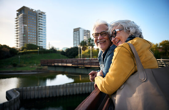 Smiling senior couple leaning on a railing in a city park, enjoying the view of a pond and cityscape, embracing their golden years together
