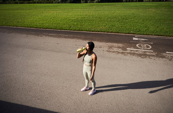 Black Latina athlete drinking from a reusable water bottle while resting on an outdoor track. High-angle fitness scene with long shadow and copy space conveying hydration and recovery.