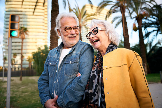 An elderly couple stands together at a crosswalk, waiting for the traffic light to change. Their quiet connection highlights care and companionship amidst the urban surroundings