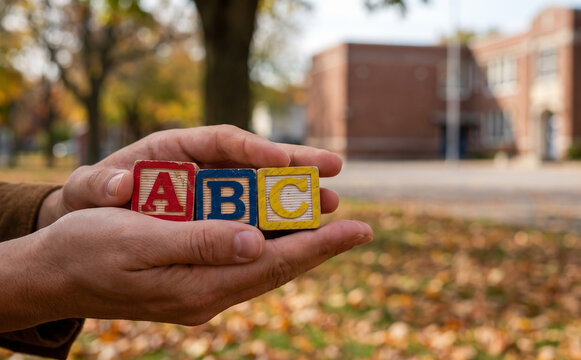 Adult hands holding ABC alphabet wooden blocks in front of a school building representing early childhood education