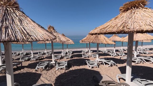 Empty resort shoreline shows rows of thatched umbrellas beside stacked lounge chairs on pebbled sand, opening to clear shallow sea with visible underwater stones and gentle wave ripples
