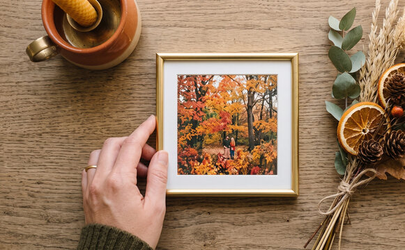 Woman holding a framed autumn photo on a wooden table with dried flowers and a candle for seasonal decor