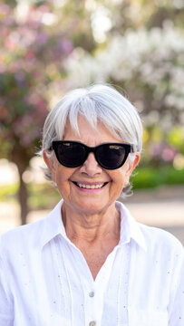 Senior woman wearing sunglasses and white shirt smiles outdoors in sunlit garden, expressing confidence and joy during relaxed summer portrait session