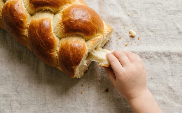 Child pulling a piece of freshly baked braided challah bread on a linen cloth