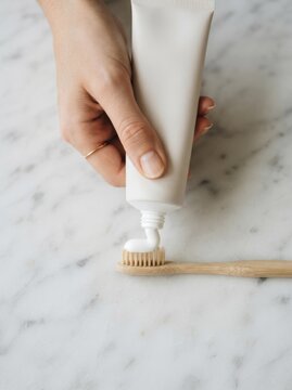 Woman squeezing organic toothpaste onto a wooden toothbrush on a marble surface, eco-friendly dental hygiene concept