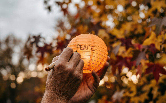Elderly hands writing the word peace on an orange paper lantern amidst autumn foliage