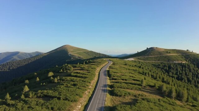 Aerial drone view of a winding mountain road passing through lush green hills and scenic countryside in warm golden light.