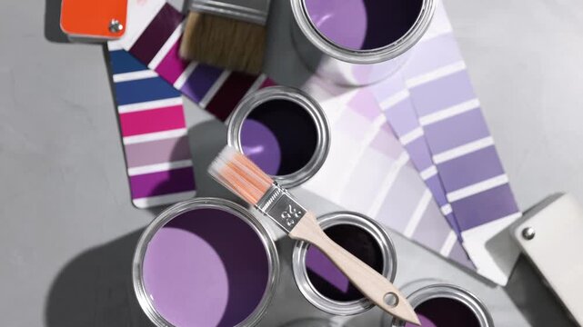 Cans with paint in shades of purple, brush and color palettes on light grey table, flat lay. Camera moving in