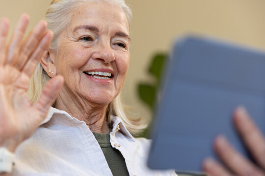Senior woman smiling and waving while holding tablet cover at home wearing light shirt, wristwatch