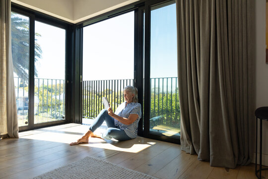 Senior woman sitting barefoot in striped shirt jeans on wood floor reading tablet near glass doors