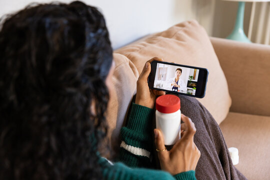 African American woman in green top on sofa holding red-capped bottle watching doctor call on phone