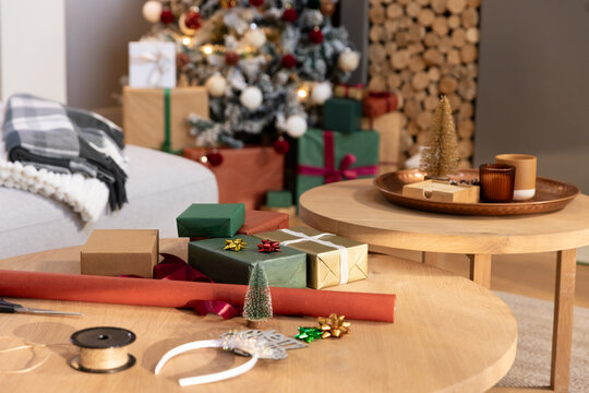 Wrapped gifts and wrapping tools are sitting on round wooden table in living room, near tree
