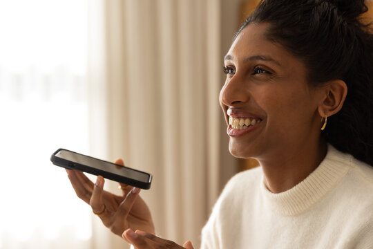 African American woman speaking into black smartphone, smiling in white sweater by window