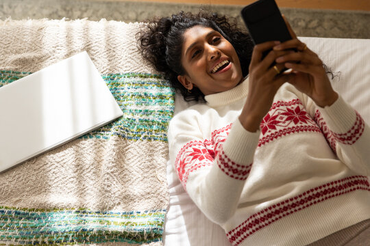 African American woman lying on bed in bedroom in white sweater holding phone beside silver laptop