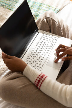 African woman sitting on bed holding silver laptop on lap, wearing cream sweater, gold rings