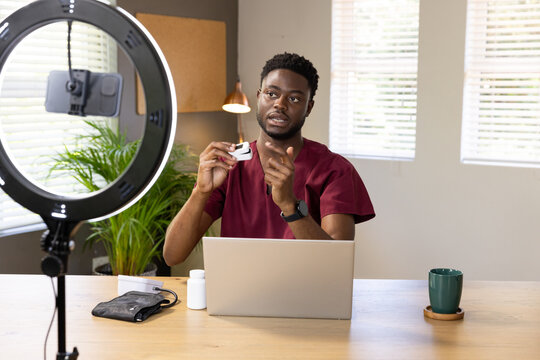 Man sitting at studio desk in maroon scrubs demonstrating device to phone on ring light