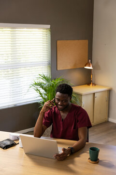 African American male wearing maroon scrubs calling on smartphone while working on laptop at office
