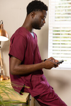 Adult African man sitting on cabinet in staff room checking phone, wearing maroon scrubs