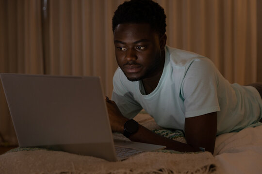 African man wearing mint T-shirt, dark watch lying on bed using silver laptop by screen light