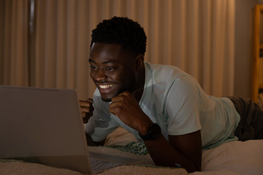 African American man lying on bed at night, facing laptop, smiling, wearing light tee, wristwatch