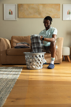 African American man folding laundry from gray basket on beige sofa with headphones, copy space
